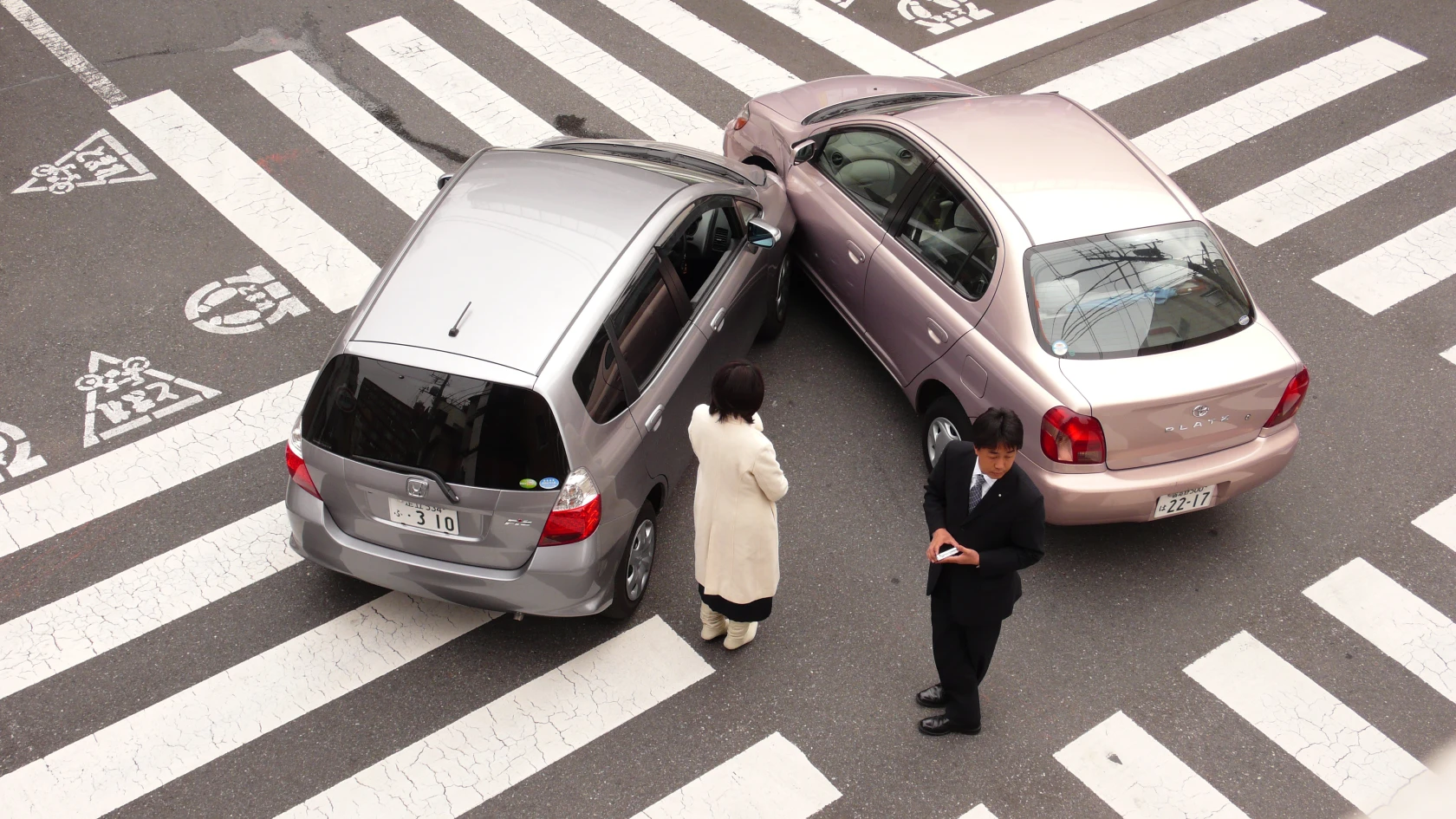 An overhead view shows two small cars involved in a minor collision in the middle of a large zebra crossing at an intersection. A silver hatchback and a pale pink sedan have bumped into each other at an angle, with visible contact between their front sides. Two people stand near the vehicles. One person in a light-colored coat stands facing the crash site, appearing still and contemplative. Another person in a dark business suit walks away while looking down at something in their hands, possibly a phone or notepad, giving the scene a detached or quietly tense feeling. The image feels calm rather than dramatic, there are no visible injuries, emergency responders, or signs of panic. The wide crossing lines and bicycle lane markings create a strong graphic pattern around the cars, adding a strangely composed, almost cinematic atmosphere to the accident scene.