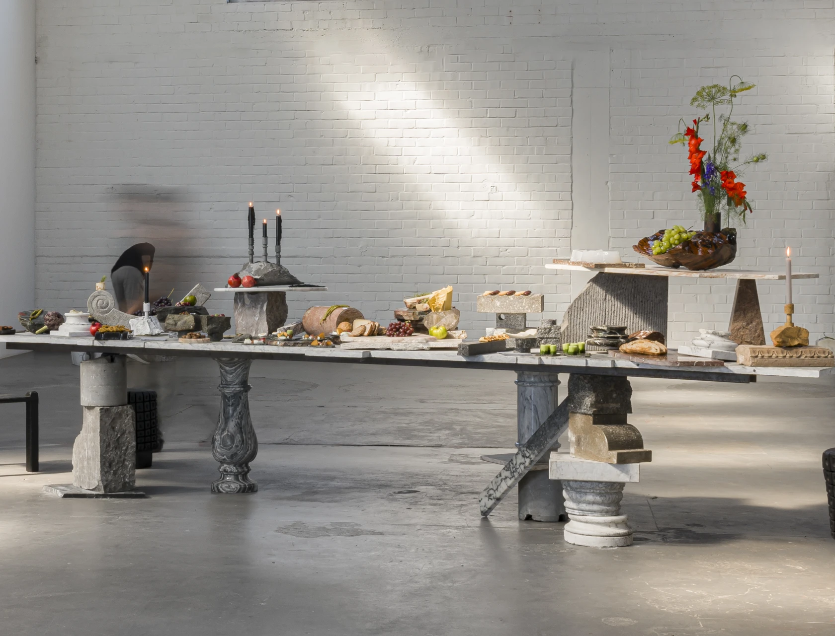 A dining table assembled from stone fragments and reclaimed architectural elements, set with food and stone tableware.