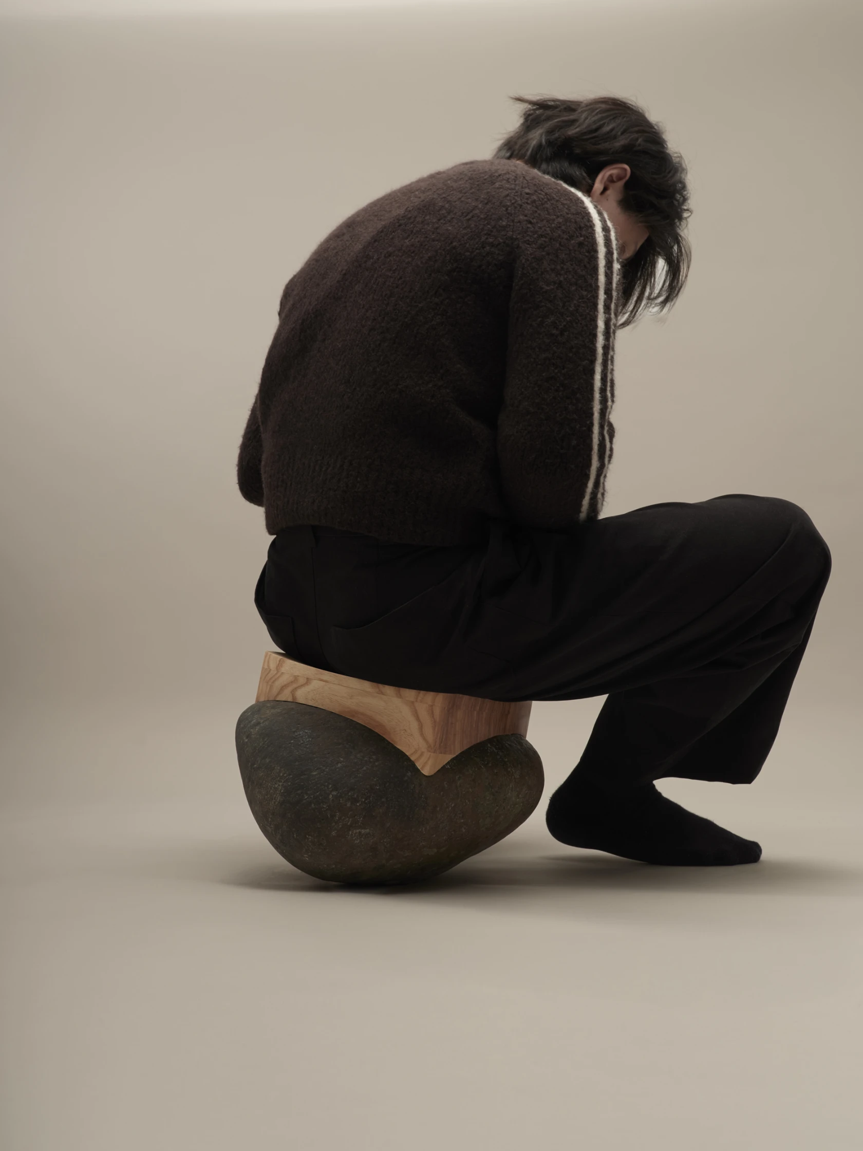 A seating object combining a natural river stone and a fitted wooden seat, with a person sitting on it, almost losing their balance.