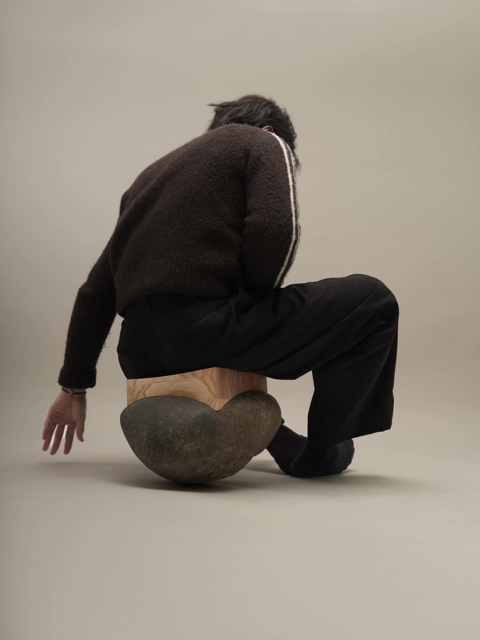 A seating object combining a natural river stone and a fitted wooden seat, with a person sitting on it, almost losing their balance.
