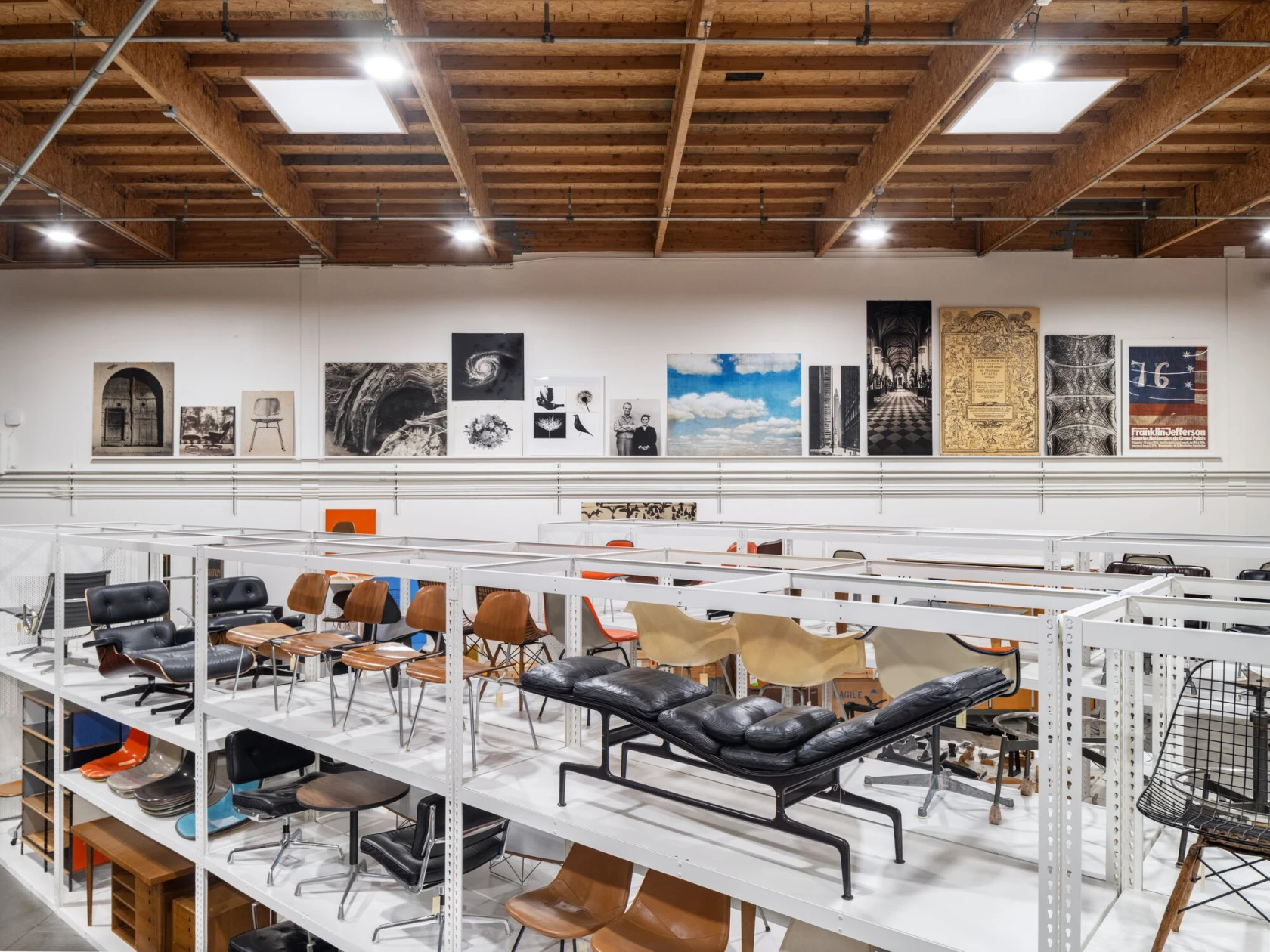 An archive space displaying a collection of furniture, objects, and visual materials, including numerous chairs and lounge pieces by Charles and Ray Eames. Framed photographs, prints, and archival materials are arranged along the wall above the open shelving system.