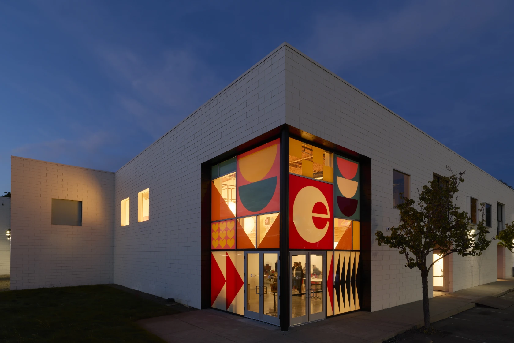 Exterior view of the Eames Institute of Infinite Curiosity at dusk, with a brightly illuminated entrance featuring colorful geometric graphics.